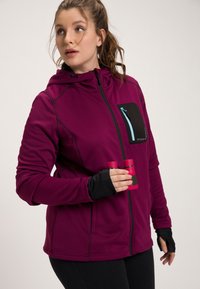 Veste d'extérieur fuchsia avec une poche poitrine zippée noire, des trous pour les pouces dans les manches et des accents noirs. Modèle tenant deux tasses roses.