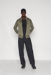 Olive roheline kinnitatav jakk tekstureeritud õlgadega, must vormitud särg, mustad kamp pants ja mustad saapad. Seismas poseerides käed puusal.