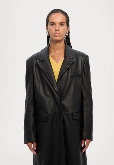 Black leather oversized blazer with a notched collar, front pocket, and button closure, layered over a yellow knitted sweater.