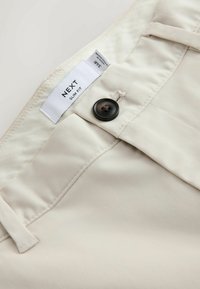 Pantalon beige slim fit avec un bouton noir et une étiquette visible à la taille indiquant la taille 34R et la marque "NEXT".