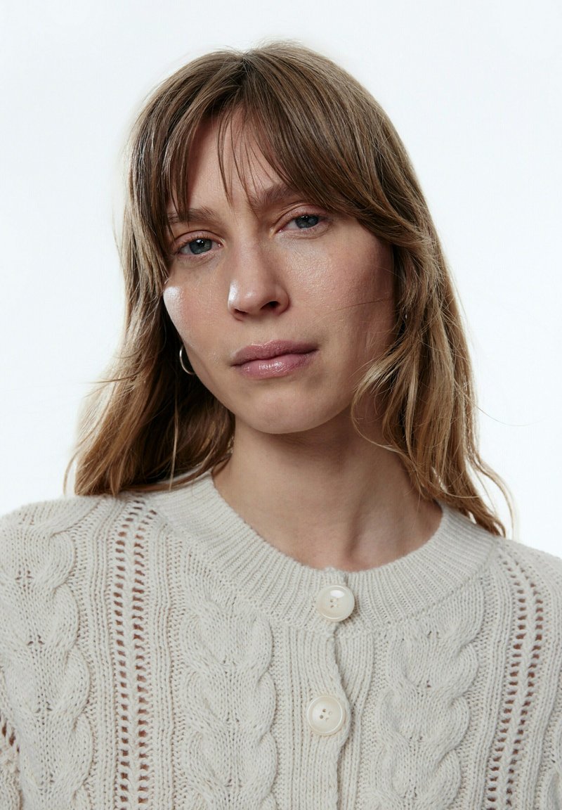 Cardigan en maille torsadée beige clair avec un col rond, fermeture à boutons et texture détaillée avec des accents de tricot ajouré.