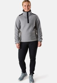Ljusgrå fleecejacka med hög krage och svarta detaljer, tillsammans med svarta byxor med dragkedjefickor och grå träningesskor.