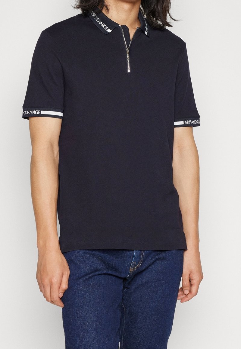 Polo-shirt marine à manches courtes, col haut et fermeture éclair. Présente des accents rayés blancs sur les manches et le col avec un logo de marque.