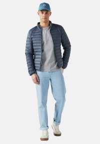 Giacca a vento da uomo in blu scuro, t-shirt grigia chiara, jeans azzurri chiari e sneaker bianche con cappellino azzurro chiaro; vestibilità comoda e design elegante.