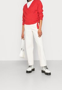 Suéter envolvente rojo de canalé con lazos, camiseta blanca, pantalones de pierna ancha blanca de tiro alto y botas blancas de cordones gruesos. Pequeño bolso blanco.