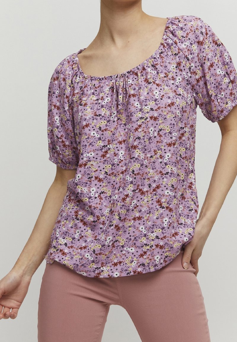 Blusa floral púrpura con mangas cortas abullonadas, escote elástico y corte holgado. Presenta una mezcla de flores blancas, amarillas y rojas en la tela.