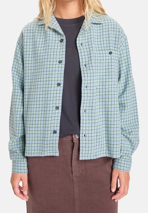 Femme portant une chemise à carreaux bleu clair et vert par-dessus un t-shirt gris foncé et un pantalon marron, debout avec les bras détendus.