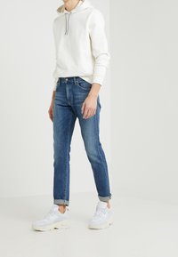 Valkoinen huppari säädettävällä hupulla, tummansiniset slim-fit farkut ja valkoiset paksut tennarit, joissa on korostuksia. Farkuissa käännetyt lahkeet.
