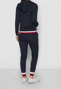 Una persona vestida con una sudadera azul marino y pantalones deportivos con puños a rayas blancas, rojas y azul marino, combinados con zapatillas blancas, de pie contra un fondo blanco.