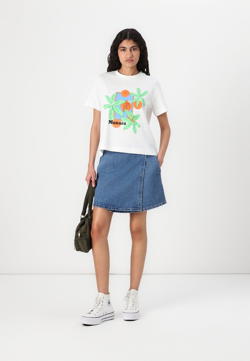 T-shirt en coton blanc avec un design floral coloré et le texte "Monaco", associé à une jupe évasée en denim bleu et des baskets montantes blanches.