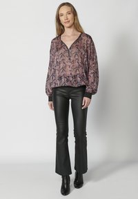 Fließende Bluse im Paisleydesign in Lila und Schwarz; V-Ausschnitt mit schwarzer Einfassung. Kombiniert mit eng anliegenden, schwarzem Schlaghosen und Stiefeletten mit Absatz.