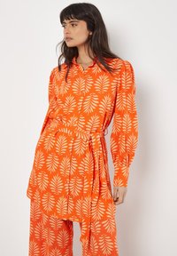 Orangefarbenes Kleid mit Blattmuster, Knopfverschluss vorne, mit umgeschlagenen Ärmeln und taillierter Taille mit Bindegürtel, aus leichtem Stoff gefertigt.