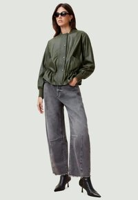 Chaqueta de cuero sintético verde oliva con textura acanalada, cierre de botones y dobladillo elástico, combinada con jeans de mezclilla grises holgados y botas negras de punta.