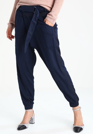 Vrouw draagt marineblauwe joggerbroek met ceintuur en elastische manchetten, gecombineerd met een lichtroze trui en zwart-witte blokhakken.