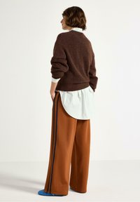 Donna che indossa un maglione marrone peloso sopra una camicia a righe con pantaloni larghi color ruggine caratterizzati da strisce scure ai lati, in piedi di lato con le mani nelle tasche.