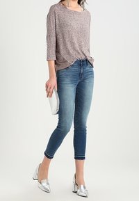 Suéter texturizado marrón claro, jeans skinny azules, zapatos de tacón plateados y un clutch plateado. Conjunto elegante y casual con acentos modernos.