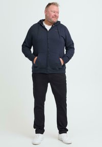 Mørk blå hettejakke med glidelås, frontlomme og snøring i hetten, kombinert med svarte bukser og hvite joggesko. Enkel, avslappet design.