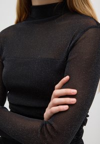 Top nero a maniche lunghe realizzato in tessuto trasparente con filo metallico, collo alto, forma aderente e dettagli testurizzati sulle maniche.