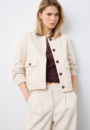 Frau trägt eine beige, geknöpfte Jacke und passende hochtaillierte Hose mit Händen in den Taschen, kombiniert mit einem dunkelbraunen, kurzen Oberteil.