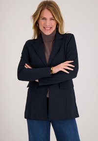 Marineblauwe blazer met een getailleerd ontwerp, grote revers en voorzakken, gedragen over een bruine coltrui. Soepele textuur, moderne snit.