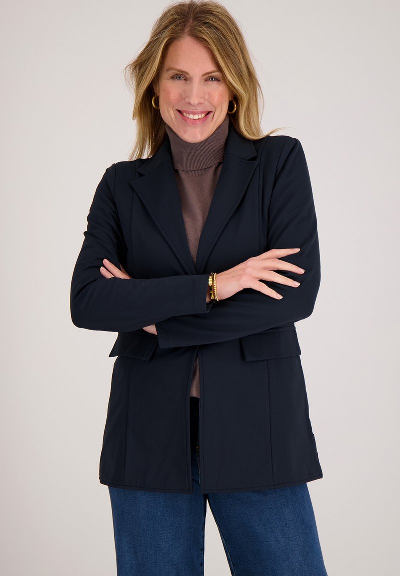 Marineblauwe blazer met een getailleerd ontwerp, grote revers en voorzakken, gedragen over een bruine coltrui. Soepele textuur, moderne snit.