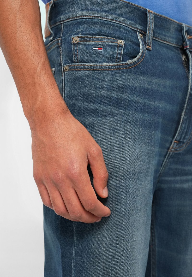 Blue denim jeans with a faded texture, showcasing a classic five-pocket design, metal hardware, and a small logo tag on the back pocket.