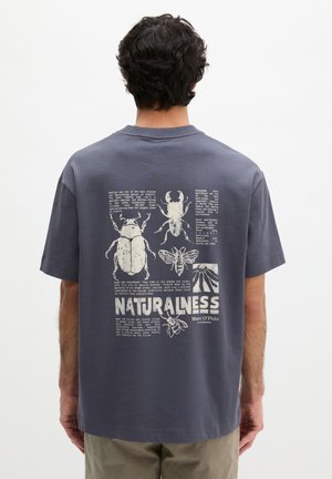 Mann trägt dunkelgraues T-Shirt mit weißen Insektenillustrationen und Text über Käfer und Natur auf dem Rücken, steht vor einem schlichten Hintergrund.