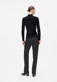 Donna con i capelli raccolti in un bun, indossa un top lungo a maniche nere sparkling, pantaloni neri dritti e stivaletti neri, in piedi di spalle su uno sfondo bianco.