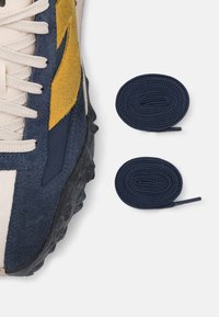 Zapato de gamuza azul y tela beige con acentos amarillos, que presenta una suela texturizada y cordones de color claro. Dos cordones de navy enrollados cerca.