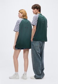 Δύο μοντέλα, γυρισμένα με την πλάτη, φορούν ταιριαστά πράσινα πουκάμισα με μοτίβο στα μανίκια και το κείμενο "EST. 1978" στην πλάτη, συνδυασμένα με casual παντελόνια.