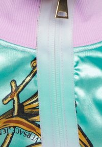 Jacka med dragkedja i ljusblå satin, med en lila ribbad krage och en detaljerad grafisk design av en kompass.