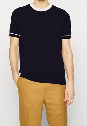 Mężczyzna ubrany w czarną dzianinową koszulkę z krótkim rękawem z białym obszyciem na kołnierzu i rękawach, w połączeniu z musztardowymi spodniami, stoi z rozluźnionymi rękami.