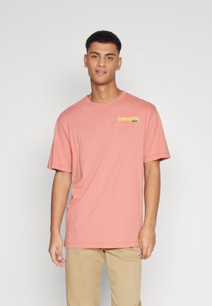 Camiseta de algodón rosa, de manga corta, con cuello redondo, que presenta un logo amarillo en el lado izquierdo del pecho. Textura suave con un ajuste relajado.
