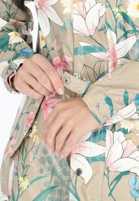 Blommmönstrad regnjacka i beige med rosa och blå blommor, med tryckknappar och en slät, glänsande yta. Detaljerade muddar synliga.