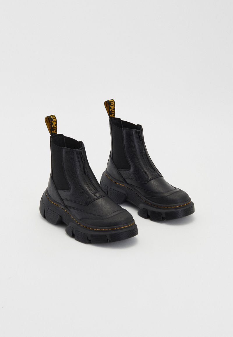 Paire de bottes en cuir noir épaisses avec semelles épaisses, fermetures éclair frontales, panneaux latéraux élastiques et détails de coutures jaunes.