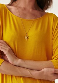 Goldene Halskette mit Katzenmotiv, aufgehängt an einer zarten Kette, getragen über einem leuchtend gelben, strukturierten Oversize-Oberteil.