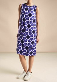 Bezramenné purpurové šaty s geometrickým vzorem v světle purpurové a černé. Hladký textil se staženým výstřihem, v kombinaci s bílými teniskami.