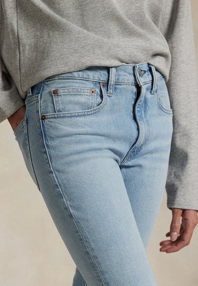 Asmuo vilki šviesiai mėlynas džinsus ir pilką ilgomis rankovėmis palaidinę, viena ranka kišenėje, kita laisvai šalia.