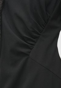 Musta mekko, jossa on teksturoitu, kerrostettu yläosa vinolla saumaerottelulla, valmistettu sileästä kankaasta. Koristeltu tyylikkäällä yksityiskohdalla.