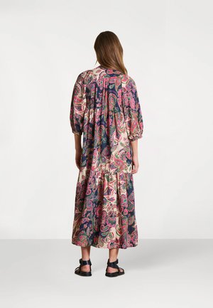 Femme debout, portant une longue robe ample avec des motifs cachemire roses et verts, ainsi que des sandales noires à bride cheville, tournée dos à la caméra.