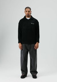 Polaire noire à col, avec un logo blanc sur la poitrine, associée à un jean gris foncé ample et des bottes noires.