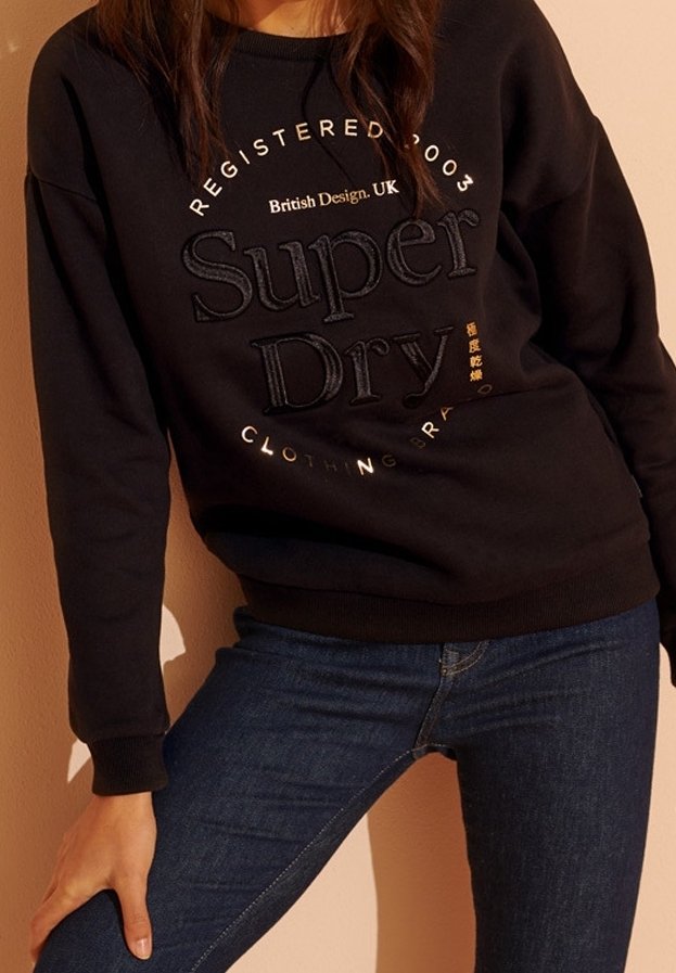 Personne portant un sweat-shirt noir Superdry avec un texte de logo brodé et un jean bleu foncé, posant devant un fond uni.
