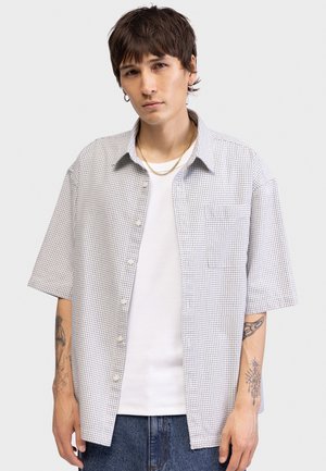 Νεαρό άτομο με κοντά σκούρα μαλλιά, φορείς κρίκους σκουλαρίκια, μια χρυσή αλυσίδα, ένα λευκό καρό κοντομάνικο πουκάμισο πάνω από λευκό τοπ και μπλε τζιν.