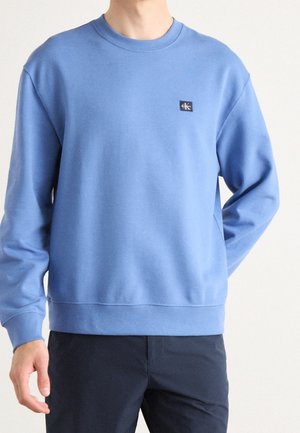 Mężczyzna ubrany w jasnoniebieską bluzę typu crewneck z małym logo CK na piersi, w połączeniu z ciemnoniebieskimi spodniami, na jednolitym jasnym tle.