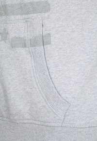 Světle šedá látka s žebrovaným okrajem kapsy a slabými tmavšími šedými skvrnami blízko levého horního rohu.