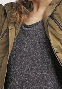 Olivově zelená zipová bunda s několika kapsami přes tmavě šedé tričko s jemným melírem, které má malou náprsní kapsu. Hladké textury po celém oděvu.
