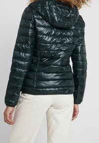 Dunkelfärgad vadderad jacka i grönt med en glänsande yta, har huva, quiltat mönster och elastiska ärmslut. Stilen kompletteras med ljusa byxor.