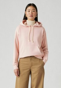 Pink hooded sweatshirt with drawstrings and a front pocket, paired with tan wide-leg trousers. Smooth fabric texture, casual design.