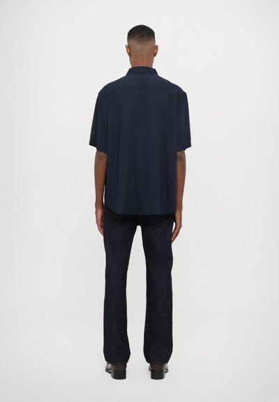 Marineblå kortermet skjorte med en lett, glatt tekstur; har krage og en enkelt knapp bak. Båret med mørke jeans og sko.