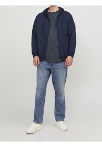 Felpa navy con zip e coulisse, indossata sopra una maglietta verde scuro. Abbinata a jeans blu sbiaditi e sneakers bianche. Design semplice.